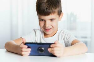 uma pequeno Garoto dentro uma branco camiseta tocam jogos em a telefone às lar. uma feliz criança parece às dele Smartphone. foto