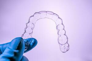 invisível dental suspensórios estão mantido de uma mão dentro uma azul luva em uma roxa fundo. plástico suspensórios odontologia retentores para endireitar dentes. foto