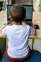 pequeno Garoto sentado às mesa usando computador portátil para conectados classe dentro grau 1, criança estudando em computador portátil a partir de casa para distância Aprendendo conectados Educação, escola Garoto crianças estilo de vida conceito foto