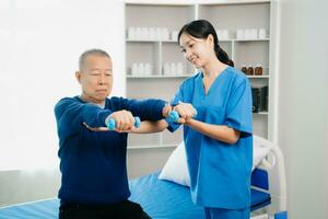 ásia fisioterapeuta ajudando idosos homem paciente alongamento braço durante exercício corrigir com haltere dentro mão durante Treinamento mão com paciente costas problemas foto