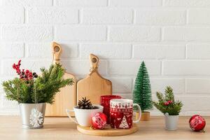 vermelho canecas com Natal padronizar em de madeira bandeja entre verde decorações com abeto galhos em cozinha feriado bancada. acolhedor lar. foto