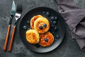 bolinhos de queijo cottage com mirtilos. cheesecakes de queijo coalho, panquecas em um fundo escuro de concreto. copie o espaço. foto