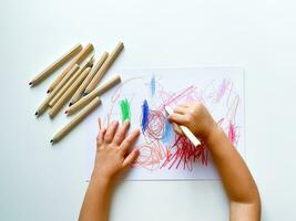 pequeno criança desenha com colori lápis em papel em branco mesa. foto