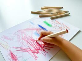 pequeno criança desenha com colori lápis em papel em branco mesa. foto