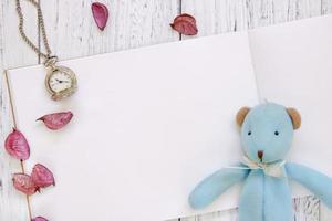 mesa de madeira pintada de branco, pétalas de flores roxas, urso, boneca, relógio de bolso