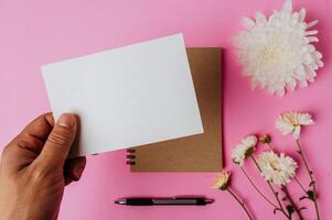 mão segurando um cartão em branco, caderno, caneta e flor em fundo rosa foto