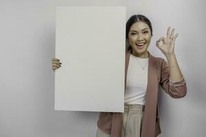 sorridente jovem ásia mulher empregado segurando e mostrando esvaziar em branco borda e gesticulando Está bem placa com dela dedos, isolado de branco fundo foto