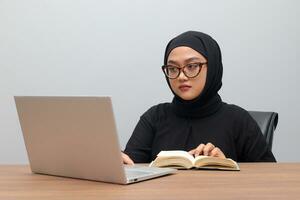 retrato do atraente ásia hijab mulher trabalhando em dela computador portátil. muçulmano menina lendo uma livro dentro escritório. empregado e freelance trabalhador conceito. foto