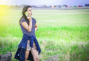 jovem menina chamando telefone em a verde campo, lindo jovem mulher chamando de célula telefone em a Relva ar livre. foto