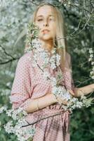 Loiras menina em uma Primavera andar dentro a jardim com cereja flores. fêmea retrato, fechar-se. uma menina dentro uma Rosa polca ponto vestir. foto