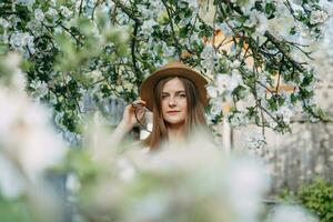 lindo jovem menina dentro branco vestir e chapéu dentro florescendo maçã Pomar. florescendo maçã árvores com branco flores foto