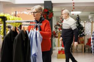 idosos mulher comprando apresenta para festivo Natal jantar, olhando para presentes dentro compras Shopping durante vendas temporada. Senior pessoa procurando para roupas em prateleiras, verificação tecidos. foto