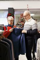 Senior pessoas olhando para blazers em prateleiras, comprando roupas em desconto a partir de roupas loja às compras Centro. clientes verificação na moda Unid, procurando festivo vestuário e presentes para Natal. foto