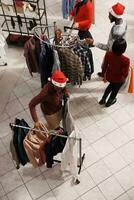 varejo escriturário organizando mercadoria em titulares dentro compras Centro loja, ajudando clientes com achando certo Natal presentes às shopping center. mulher loja empregado comovente cabides do Itens em volta. foto