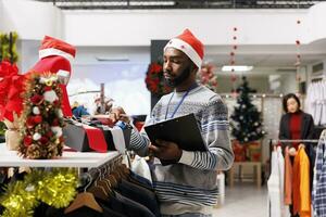 homem loja Gerente contando laços, criando Lista do mercadoria estoque para inventário dentro roupas loja. africano americano varejo empregado arranjo produtos e moda Itens às compras Centro. foto