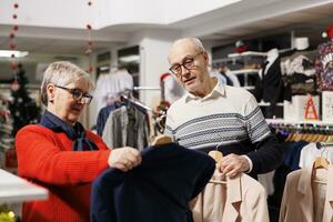 dois clientes examinando terno jaquetas dentro roupas loja, revendo blazers em cabides e visitando Shopping para Comprar apresenta. homem e mulher tentando para escolher 1 roupas item em desconto, natal temporada. foto