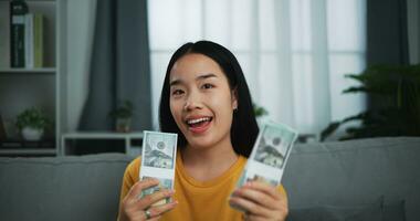 retrato do jovem ásia mulher mostrando dólar às Câmera e sorridente em sofá dentro a vivo quarto às lar. foto