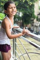 pequeno criança jogando dentro verão chuva dentro casa sacada, indiano inteligente Garoto jogando com chuva gotas durante monção chuvoso temporada, criança jogando dentro chuva foto