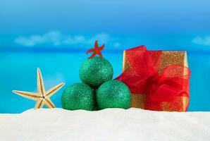 verde brilhante Natal árvore fez do brilhar bolas com estrela do Mar, embrulhado presente caixa com vermelho arco em areia do praia, mar atrás. Novo ano, feriados dentro quente países foto