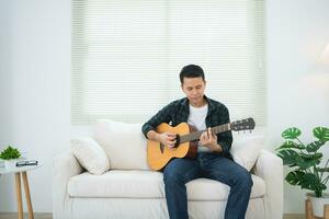 ásia homem jogando guitarra sentado em sofá sofá ou chão dentro a vivo quarto às lar. ásia homem escrevendo música enquanto jogando guitarra às lar. compor música música conceito. foto