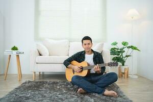 ásia homem jogando guitarra sentado em sofá sofá ou chão dentro a vivo quarto às lar. ásia homem escrevendo música enquanto jogando guitarra às lar. compor música música conceito. foto