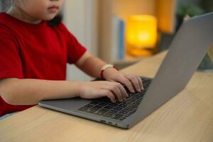 fechar acima do mão bebê, ásia bebê menina vestindo uma vermelho camiseta usar computador portátil e estude conectados em madeira mesa escrivaninha dentro vivo quarto às lar. Educação Aprendendo conectados a partir de casa conceito. foto