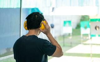 homem atirador asiático usando fones de ouvido com cancelamento de ruído e roupas pretas praticando tiro de arma curta no campo de tiro. tiro esportivo para meditação e autodefesa, atividades recreativas. foto