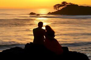 caucasiano casal sentado em Rocha perto mar às pôr do sol foto