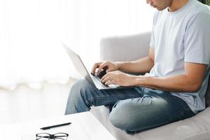 jovem sentado no sofá, digitando no laptop para trabalhar online. foto