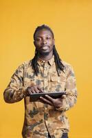 africano americano homem segurando tábua computador, testa em Internet dentro estúdio sobre amarelo fundo. sorridente adulto rolagem cocho social meios de comunicação, assistindo engraçado vídeo em digital dispositivo foto