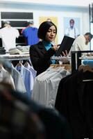 roupas loja ásia mulher empregado usando digital tábua enquanto trabalhando dentro compras Centro. departamento Shopping boutique trabalhador examinando vestuário dentro estoque e gerenciando inventário foto