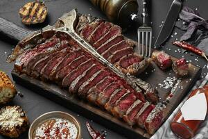 fatiado bife t-bone deitado em de madeira borda. grelhado legumes dentro uma panela grade. Preto mesa com cinzento pano foto