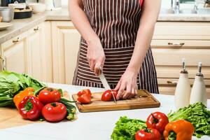 jovem mulher cozinhando dentro a cozinha às lar. uma mulher cortes uma tomate e legumes com uma faca. foto