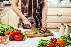 jovem mulher cozinhando dentro a cozinha às lar. uma mulher cortes uma pepino e legumes com uma faca. foto