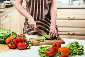 jovem mulher cozinhando dentro a cozinha às lar. uma mulher cortes uma pepino e legumes com uma faca. foto