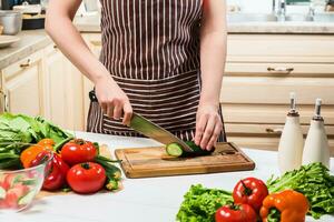 jovem mulher cozinhando dentro a cozinha às lar. uma mulher cortes uma pepino e legumes com uma faca. foto