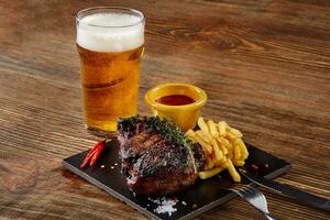 cerveja sendo derramada em vidro com bife gourmet e batatas fritas em fundo de madeira. foto