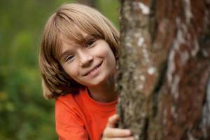 menino de camiseta vermelha espiando por trás de um tronco de árvore foto