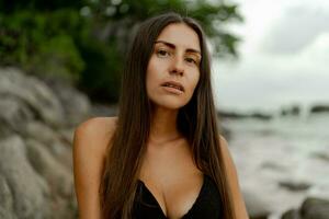 retrato do morena mulher com perfeito figura posando em tropical rochoso de praia. vestindo à moda Preto roupa de banho. foto