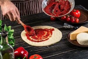 uma masculino mão espalhando tomate purê em uma pizza base com colher em a velho de madeira fundo foto