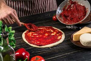 uma masculino mão espalhando tomate purê em uma pizza base com colher em a velho de madeira fundo foto