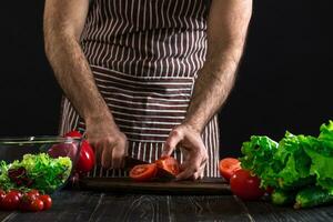 homem preparando salada em uma de madeira mesa. masculino mãos cortar a tomate para faço uma salada em Preto fundo foto