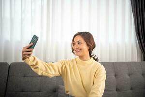 a ásia menina com uma grande sorrir senta dentro a vivo sala. ela tem Diversão levando alegre usar Smartphone vídeo conferência selfies e olhando às a Câmera foto