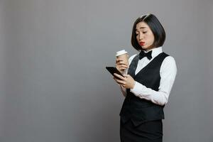 refeições serviço ásia garçonete conversando conectados usando Smartphone e bebendo levar longe café. restaurante mulher trabalhador digitando mensagem em Móvel telefone enquanto desfrutando quente bebida para ir foto