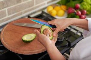 mulher mãos segurando a abacate fruta e descamação isto enquanto em pé às a cozinha contador, preparando saudável salada foto