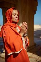 lindo árabe muçulmano mulher dentro brilhante laranja autêntico vestir e dela cabeça coberto dentro hijab, Rezar com dela mãos acima foto