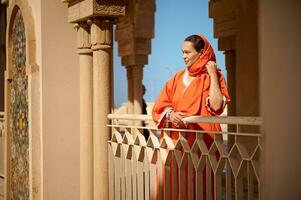 lindo mulher dentro à moda oriental vestuário e coberto cabeça dentro cachecol do laranja cor, olhando para dentro distância, dentro uma mesquita foto