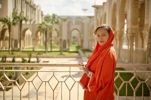 encantador muçulmano mulher dentro laranja lenço, olhando às Câmera, em pé em a sacada contra a mesquita jardim fundo foto