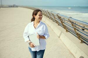 lindo feliz latim americano jovem o negócio mulher segurando computador portátil, caminhando ao longo a passeio e olhando às Beira Mar foto