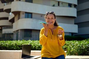 sorridente feliz mulher com computador portátil, usando inteligente Móvel telefone, gravação voz mensagem contra Alto subir construção fundo foto